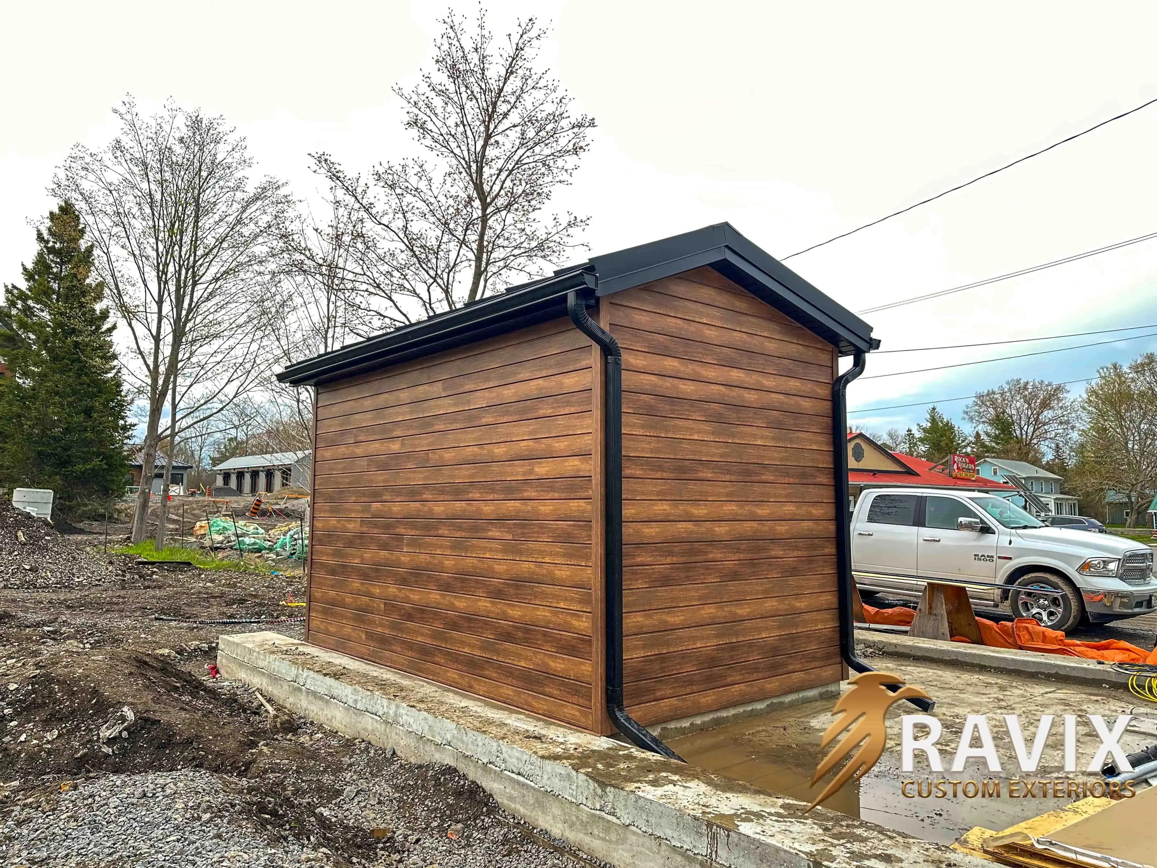 Wood-look exterior siding installed on a small utility building with black fascia, soffit, eavestrough, and downspouts.