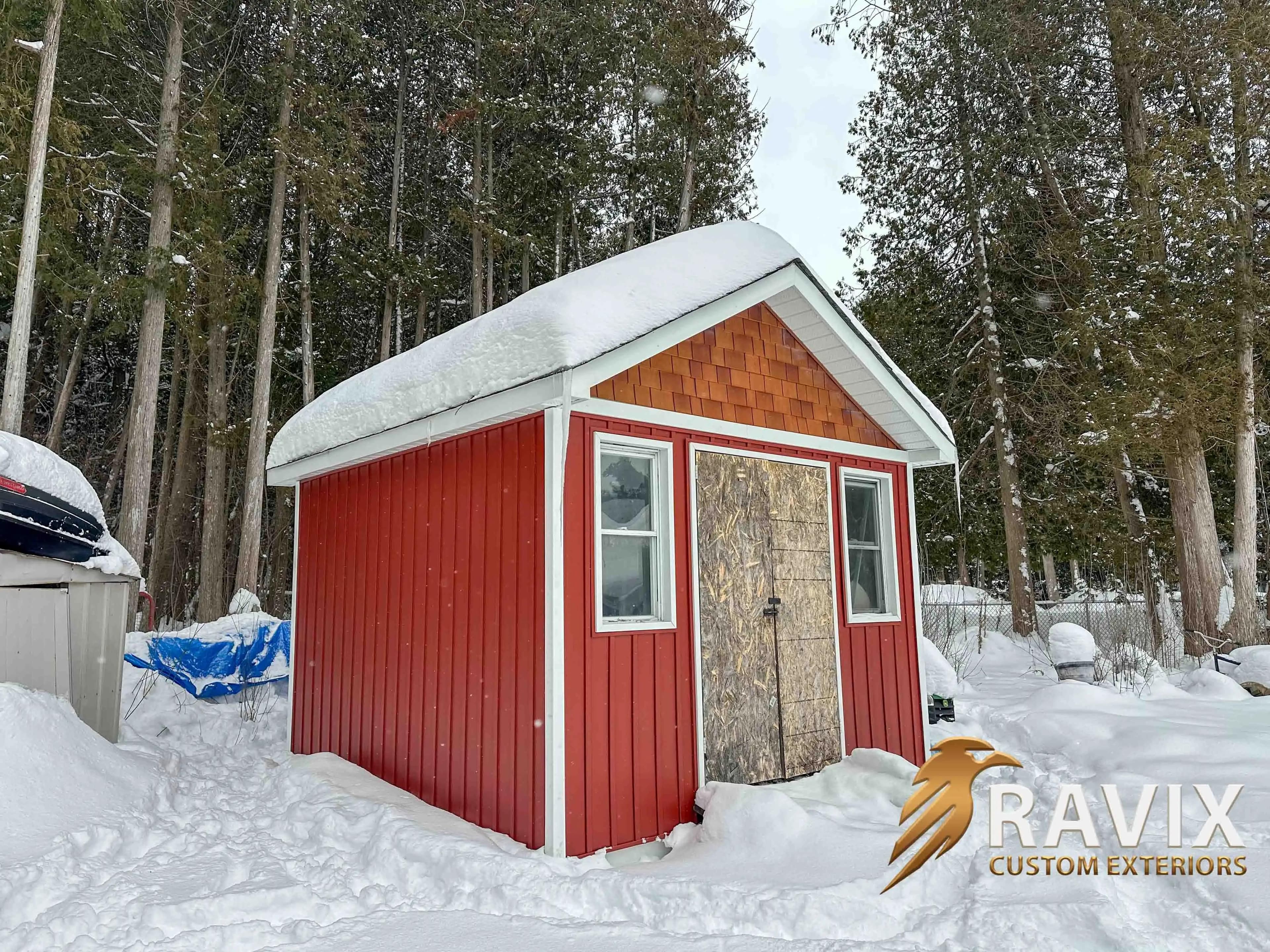 Red shed with white trim, cedar shake gable accent, windows, and a snow-covered roof in a wooded winter setting.