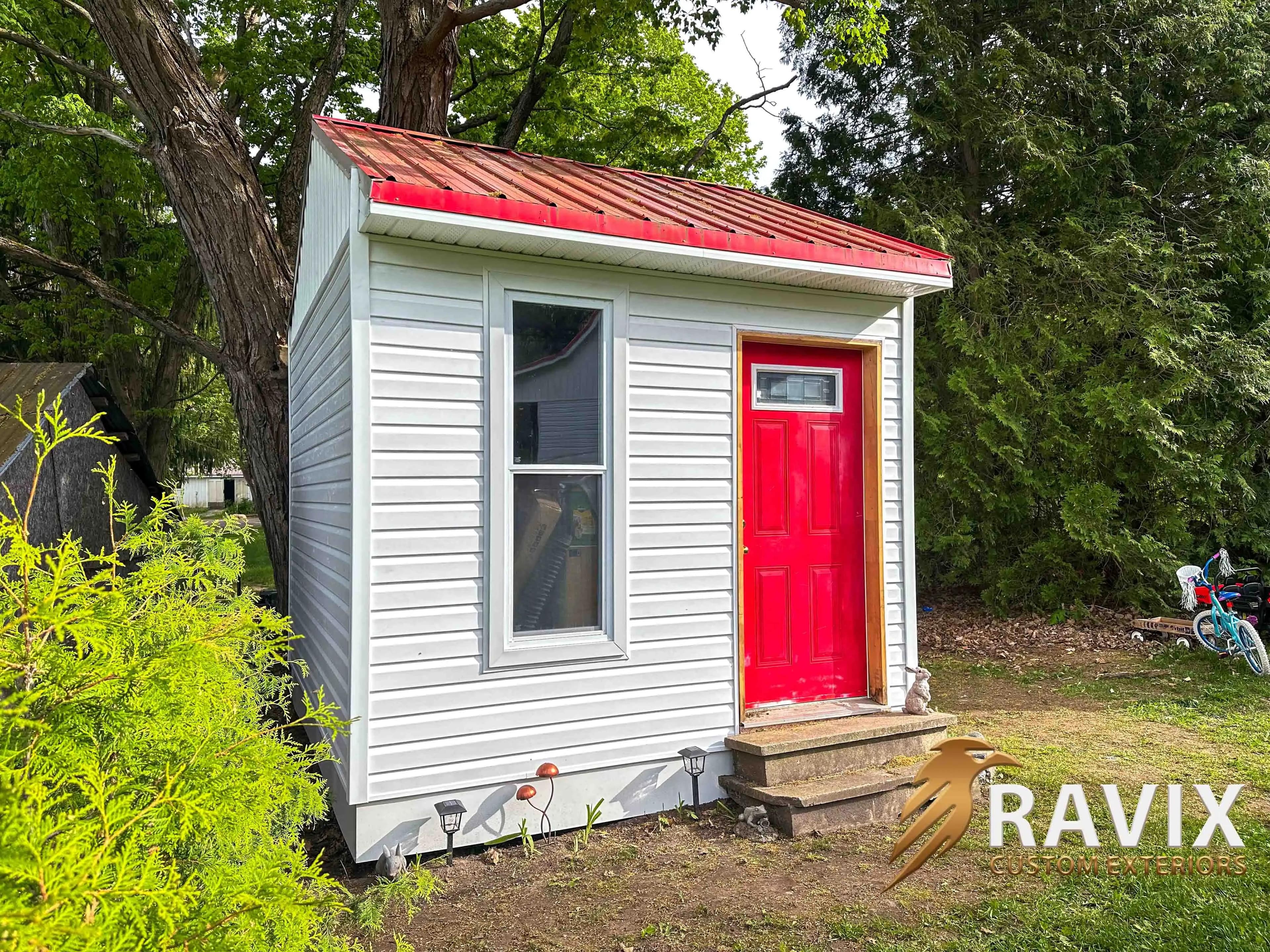 Small shed finished with white vinyl siding, a red metal roof, and a bright red front door surrounded by trees.