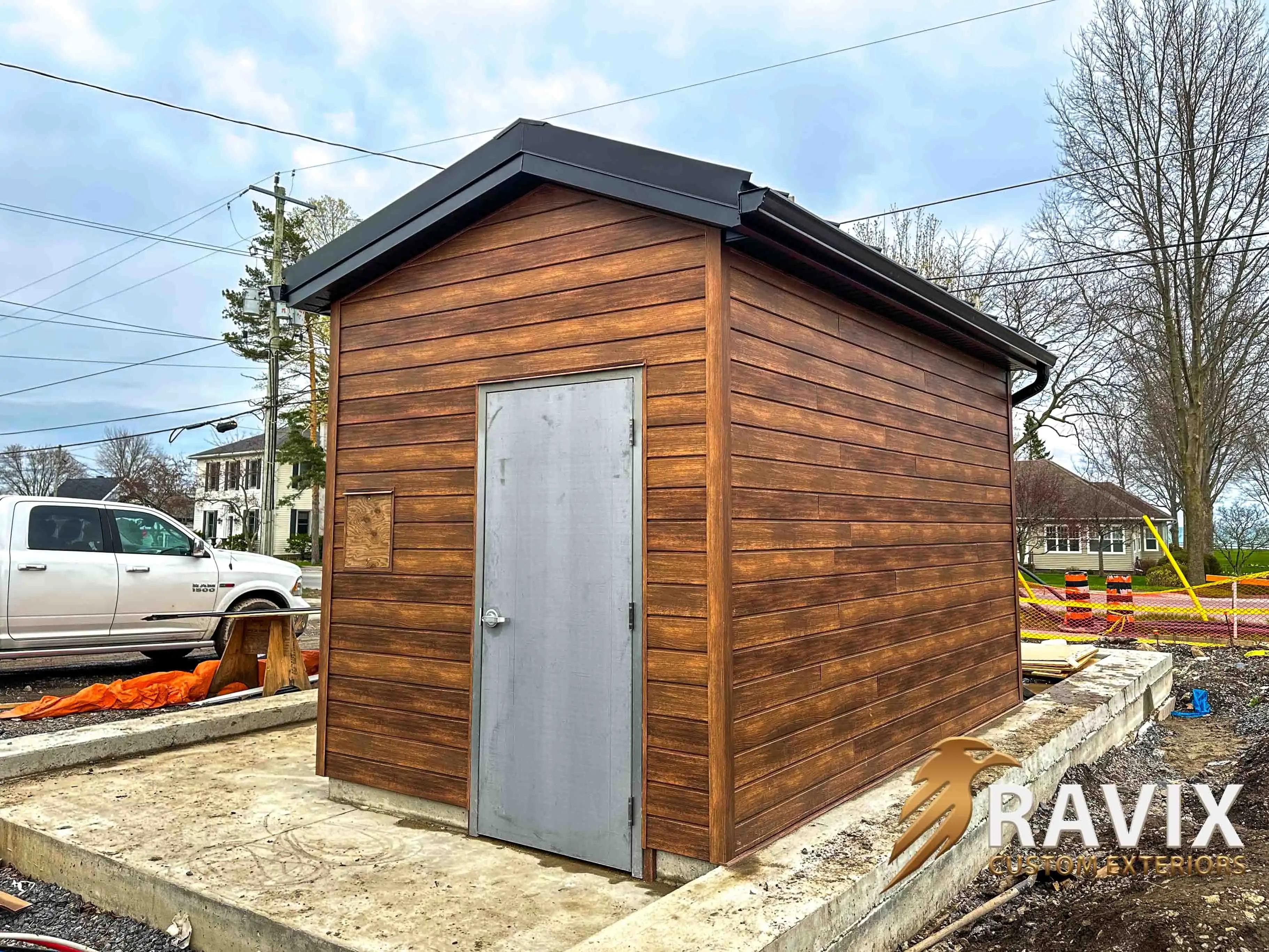 Small exterior building finished with horizontal wood-look siding, black metal trim, and a grey utility door.