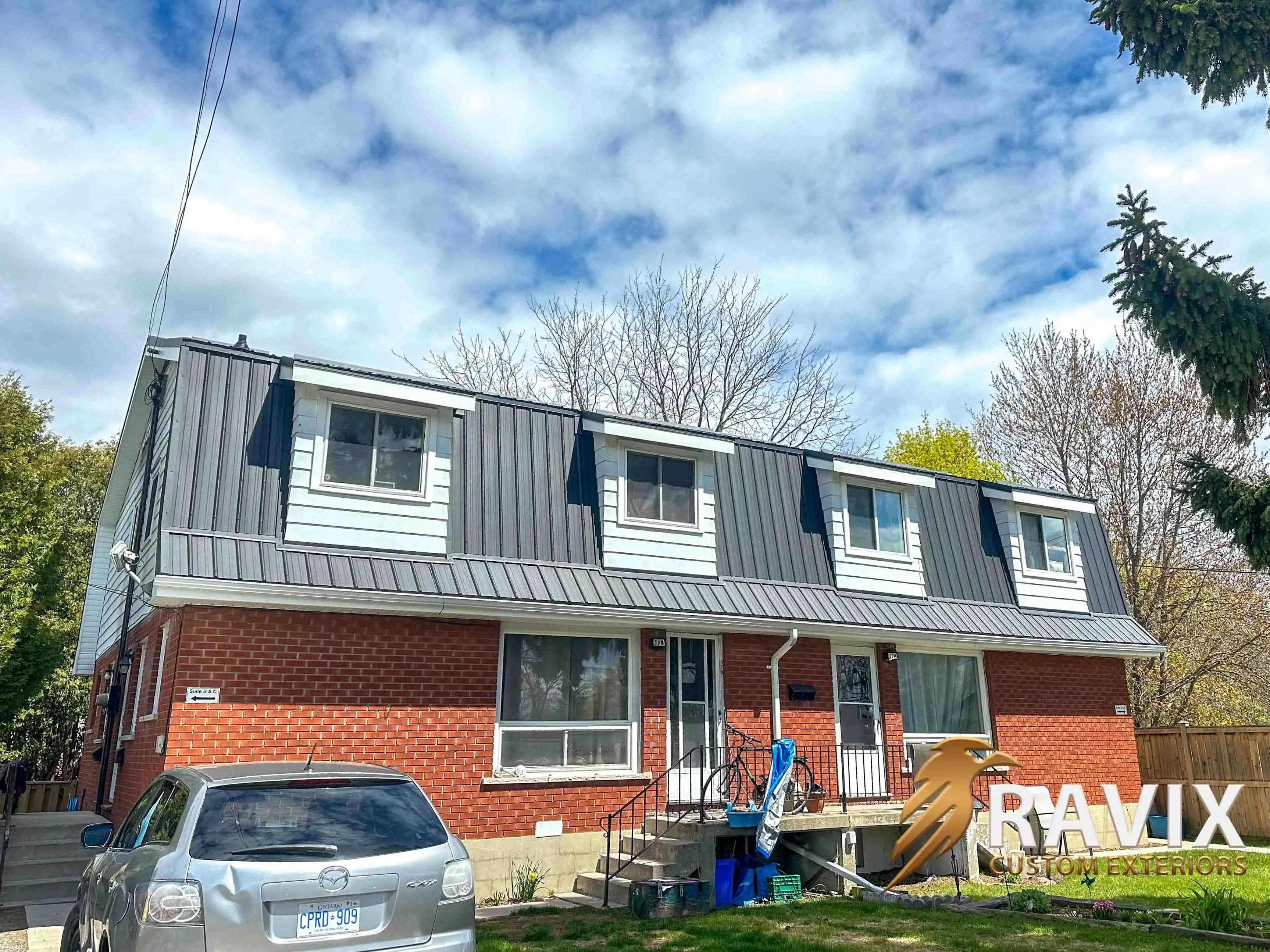 Angled view of a brick residential building with dark metal roofing, white dormers, white gutters, and updated exterior details.
