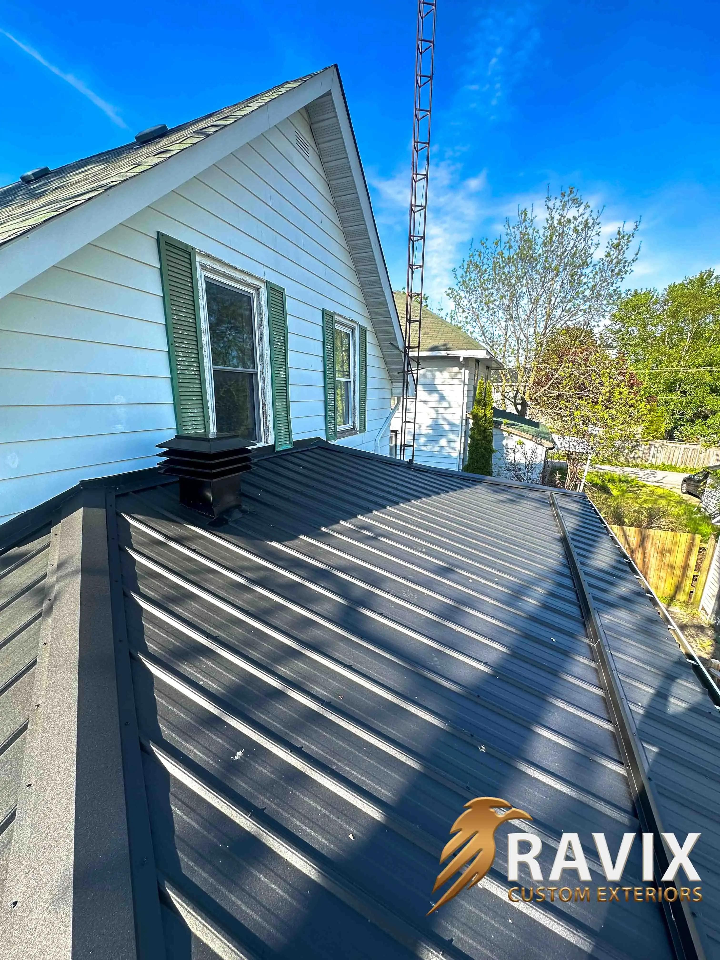 Top view of a dark metal roof with black trim, roof vent, and white siding on a sunny residential property.