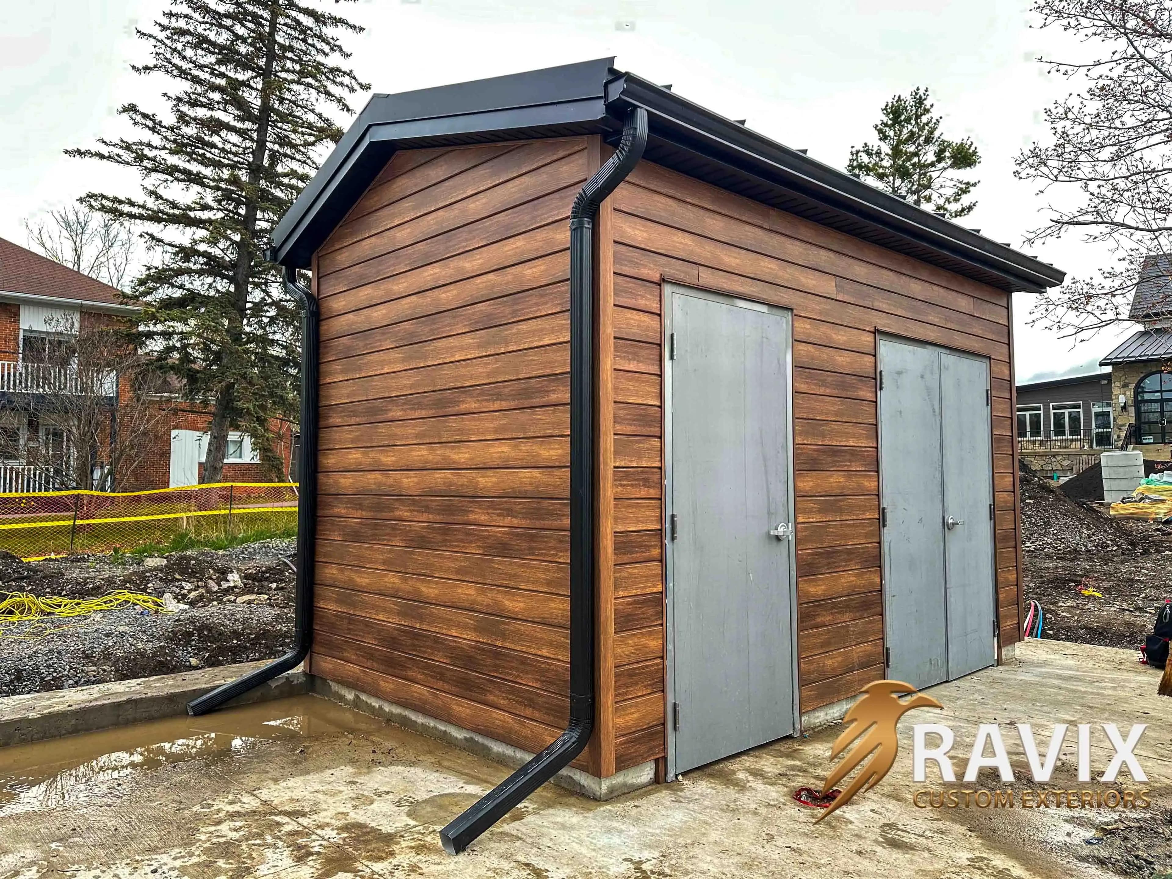 Finished small building with wood-look siding, black roof trim, black gutters, and downspouts on a construction site.