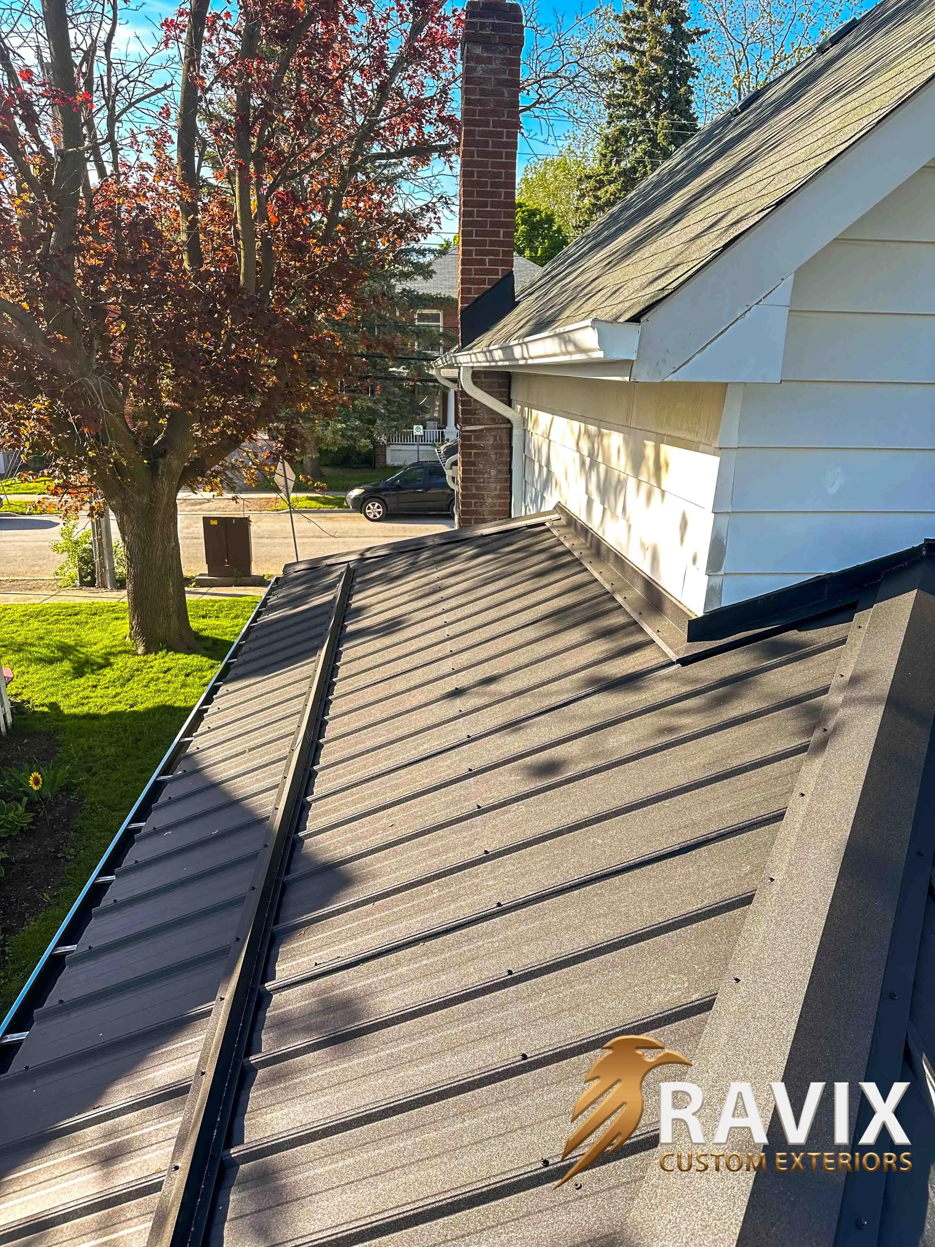Close-up view of a dark metal roof installed on a residential home with white fascia, eavestrough, and chimney flashing.