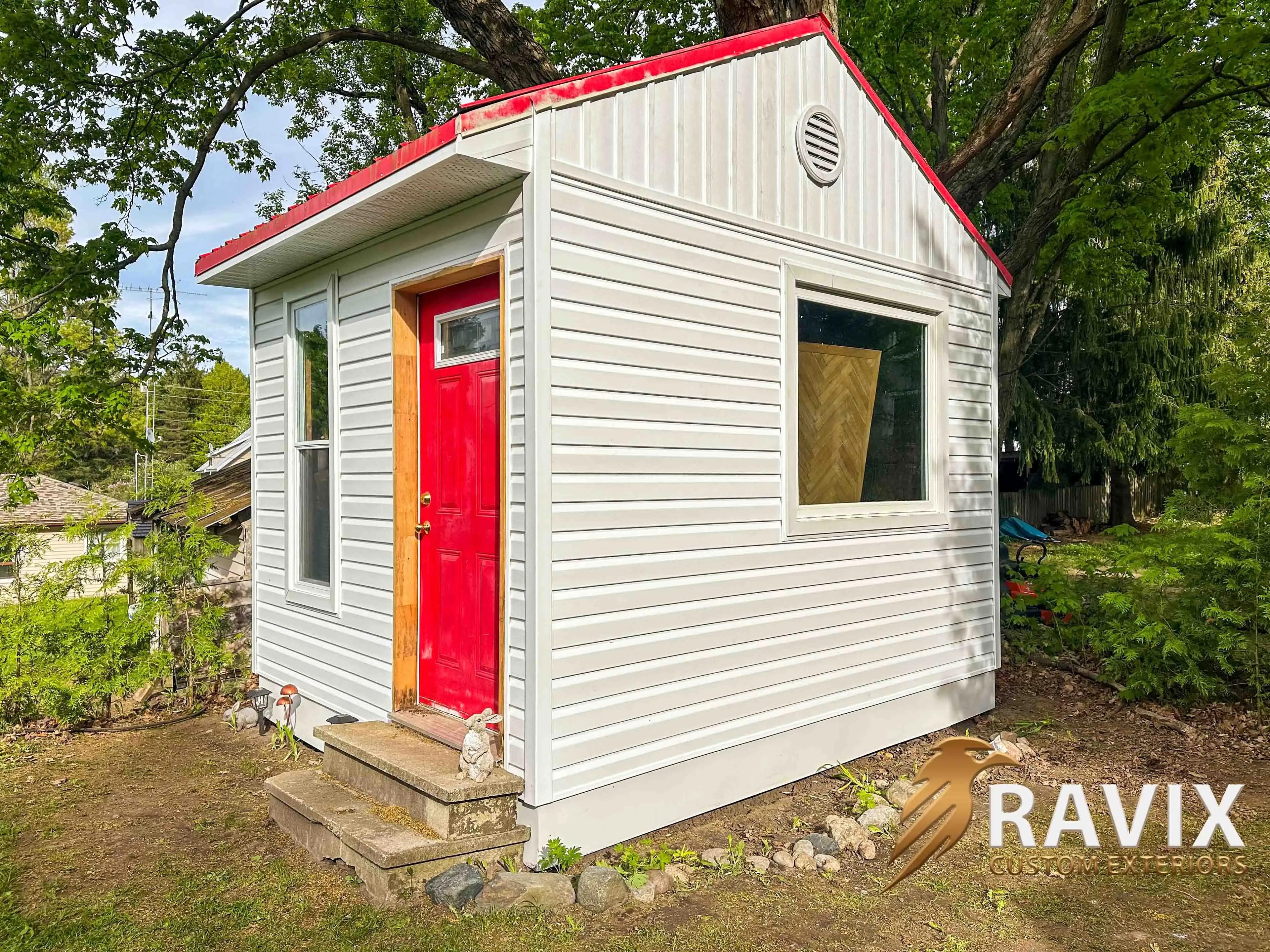 Side view of a small shed with white horizontal siding, white trim, a red door, and red roof trim.